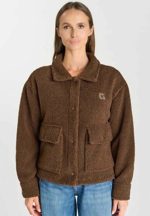 Femme portant une veste marron duveteuse avec poches boutonnées et col, associée à un jean bleu, debout devant un fond uni.