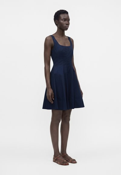 Femme debout portant une robe sans manches bleu marine longueur genou et des sandales plates marron sur un fond clair uni.