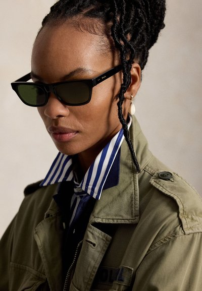 Fonott hajú nő fekete téglalap alakú napszemüveget, csíkos inget, kagylófülbevalót és olívazöld dzsekit visel, balra néz.