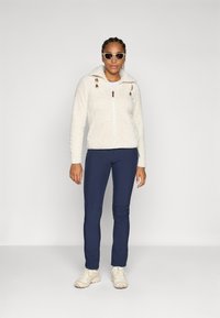 Veste blanche en polaire avec des accents marron, pantalon ajusté bleu marine et baskets claires. Modèle portant des lunettes de soleil, se tenant devant un fond uni.