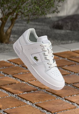 White leather sneaker with perforated toe, lace closure, and embroidered green crocodile logo. Textured sole with raised edges.