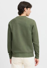 Sudadera verde oliva con puños y dobladillo acanalados. Tejido de mezcla de algodón, escote redondo y textura suave. Combinada con pantalones color crema.