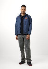 Marineblå vindjakke med lynlås foran, orange logo, sort skjorte under, grå jeans og sorte sneakers med røde detaljer.