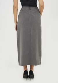 Szara, długa spódnica o slim fit, z gładką teksturą i dwiema kieszeniami z tyłu. Noszona z czarnymi butami na wysokim obcasie.
