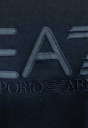 T-shirt di cotone blu navy scuro con testo ricamato in rilievo. "EA7" in grandi lettere grigie con "Emporio Armani" sotto, accompagnato da dettagli del logo.