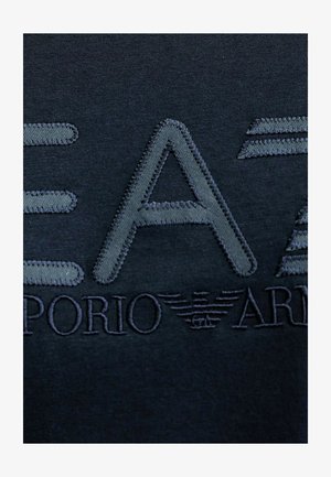 T-shirt di cotone blu navy scuro con testo ricamato in rilievo. "EA7" in grandi lettere grigie con "Emporio Armani" sotto, accompagnato da dettagli del logo.
