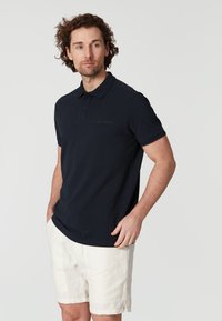 Marinblå pikétröja med klassisk krage, korta ärmar och en subtil logotypdetalj på bröstet, i kombination med ljusa shorts.