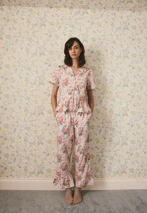 Naine seisab lillelise tapeedi taustal, kandes roosat lillemustriga öösärki, millel on volangidega ääred, käed taskutes, seljas beežid sokid.