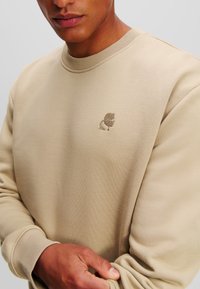 Sudadera beige con cuello redondo, puños acanalados y logo bordado en el pecho. Textura suave con un acabado liso.