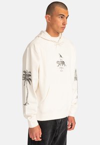 Cream hoodie featuring a central graphic of a bird and a tiger, with palm tree designs on the sleeves. Made from soft fabric, casual fit.