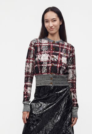 Kvinde iført en rød, sort og hvid ternet knap-sweater med grå manchetter og talje, kombineret med et skinnende sort tekstureret skørt.