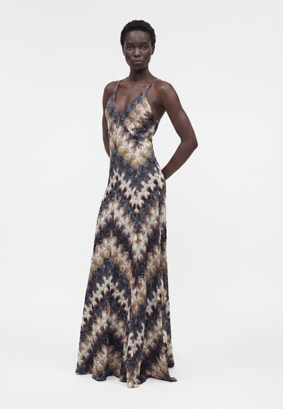 Γυναίκα στέκεται με τα χέρια πίσω από την πλάτη της, φοράει αμάνικο μάξι φόρεμα με V-λαιμόκοψη και πολύχρωμο ζιγκ ζαγκ μοτίβο.