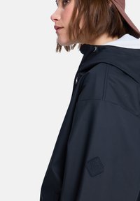 Μαύρο αδιάβροχο μπουφάν με κουκούλα, με υφή στην επιφάνεια, ραμμένες λεπτομέρειες και λογότυπο σε κεντημένη ετικέτα στο μανίκι.