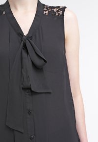 Blouse noire sans manches avec des détails en dentelle sur les épaules, un nœud noué au niveau du col et une fermeture à boutons à l'avant. Tissu lisse, coupe ample.