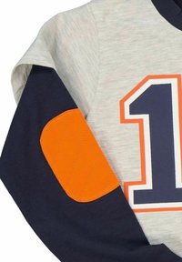 Harmaa huppari, jossa on tummansiniset hihat, oranssi tikattu kyynärpääpala ja suuri oranssi ja tummansininen numero "1" painettuna rintakehälle.