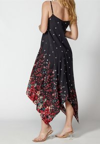 Femme portant une robe noire à ourlet mouchoir à motif floral avec fines bretelles, associée à des sandales à talons hauts transparentes, debout devant un fond uni.