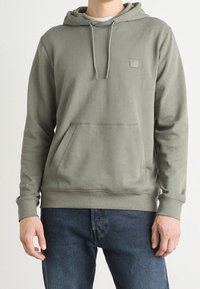 Homme portant un sweat à capuche vert clair avec une poche avant et des cordons, associé à un jean bleu en denim, debout devant un fond uni.