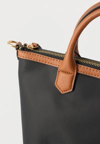 Černá textilní taška s hnědými koženými prvky, zlatým zipem a texturovanou koženou rukojetí. Obsahuje kovový kroužek připevněný k táhlu zipu.