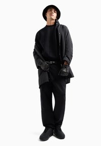 Giacca nera con alto colletto, maglione nero in tessuto strutturato, pantaloni neri e sneakers. Guanti neri e un cappello completano l'outfit.