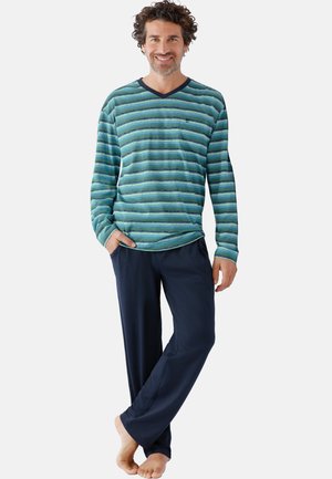 Lächelnder Mann, der ein grün-blau gestreiftes Langarmshirt und dunkelblaue Hose trägt, steht barfuß mit einer Hand in der Tasche.