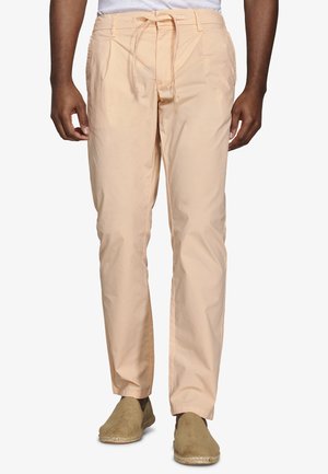 Mężczyzna ubrany w jasnoróżowe spodnie ze ściągaczem i beżowe wsuwane buty, stojący z rękami swobodnie opuszczonymi wzdłuż ciała na białym tle.