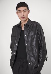 Chaqueta de camisa de cuero negro con cierre a presión, dos bolsillos en el pecho y costuras detalladas. Usada sobre una blusa negra ajustada.