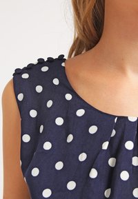 Navy sleeveless blouse with white polka dots, pleated design, and button accents on the shoulder. Soft, lightweight fabric texture.