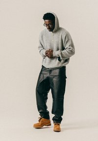 Hombre que lleva una sudadera de punto gris, pantalones de cuero negros, botas de trabajo marrones y gafas, de pie con las manos juntas y la cabeza ligeramente inclinada.