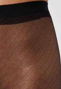 Čierny pás so šikmým diamantovým vzorom na priesvitných pančuchách v stredne hnedej farbe, s hladkou textúrou a ľahkým materiálom.