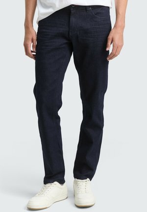 Dunkelblaue Jeans mit geradem Schnitt, ausgestattet mit vorderen Taschen, einem dezenten Verblassen und einem kontrastierenden Nähdetail, kombiniert mit weißen Sneakern.