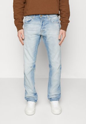 Mężczyzna ubrany w jasnoniebieskie wyblakłe jeansy, brązową bluzkę z długim rękawem i białe trampki, stoi ze swobodnie opuszczonymi rękami przy bokach.