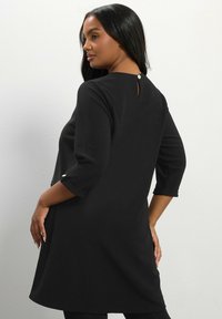 Abito tunica nero con maniche 3/4, scollatura rotonda e dettaglio a chiave sul retro con un bottone decorativo. Tessuto morbido con vestibilità ampia.