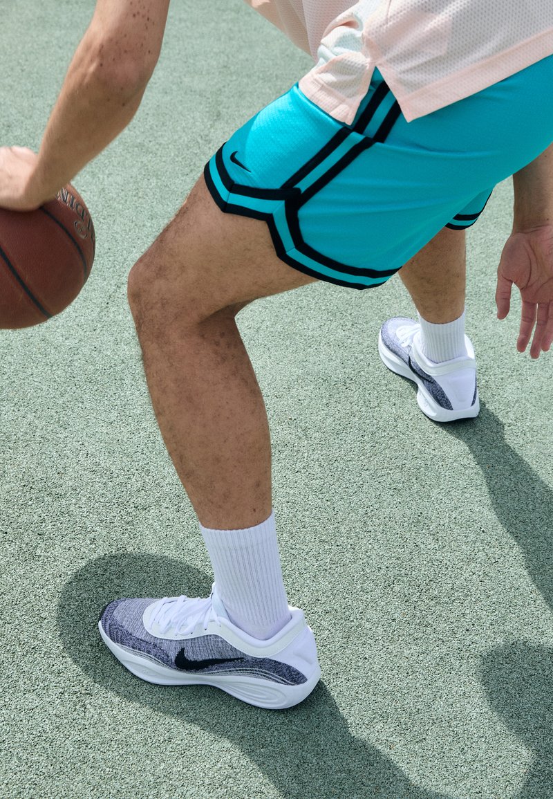 Des chaussures de sport blanches et grises avec une tige texturée sont portées avec un short teal présentant des accents noirs. L'arrière-plan montre un ballon de basket et un terrain de jeu.