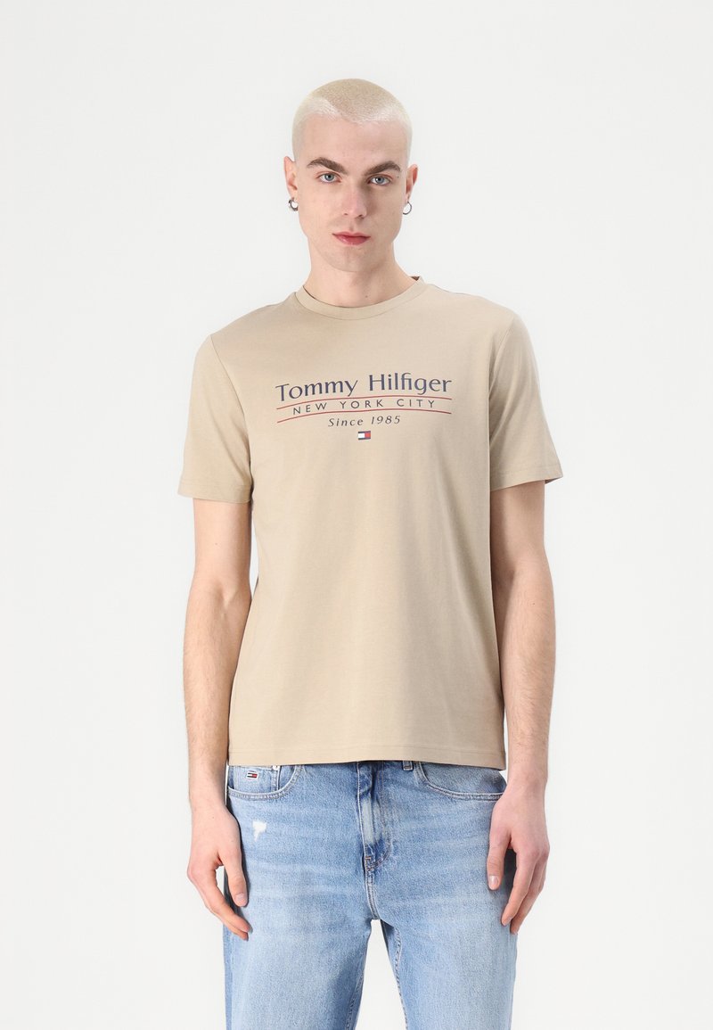 Beige kortermet t-skjorte med teksten "Tommy Hilfiger New York City Since 1985" i marineblått og rødt, med rund hals og myk bomullstekstur.