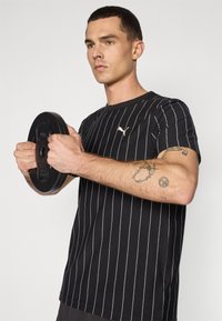 Sort T-shirt med korte ærmer og hvide lodrette striber, med et lille guld logo. Personen holder en sort vægttallerken.