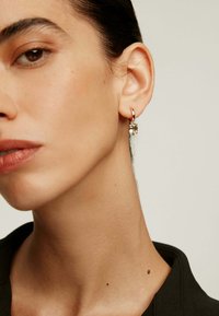 Pendiente de aro de oro con un detalle de piedra clara, con un acabado pulido. El modelo tiene la piel suave y viste una camiseta oscura.