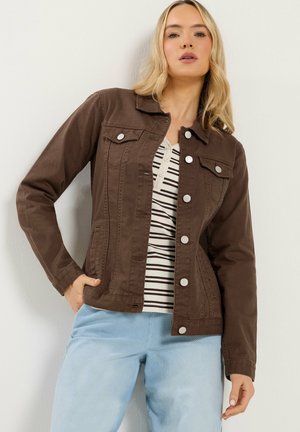 Kobieta w brązowej zapinanej kurtce na białej i czarno paskowanej koszulce, w jasnoniebieskich dżinsach, stojąca na tle białej ściany.