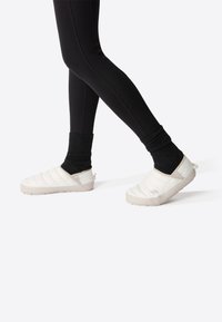 Chaussures blanches à enfiler rembourrées avec une tige texturée, une bande élastique et des semelles en caoutchouc légères, associées à des chaussettes noires côtelées et des leggings.