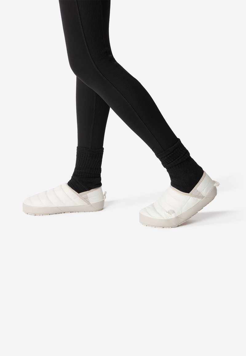 Chaussures blanches à enfiler rembourrées avec une tige texturée, une bande élastique et des semelles en caoutchouc légères, associées à des chaussettes noires côtelées et des leggings.