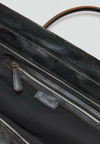 Interno di una borsa in finta pelle nera con texture, dotato di una tasca con zip, cerniera in metallo argentato e etichetta MNG. Cuciture marroni evidenziano i bordi.