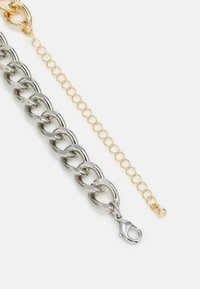Pulseira de corrente de metal prateado com fecho tipo lagosta, acompanhada de uma fina corrente extensora dourada com elos ovais e uma conta redonda.