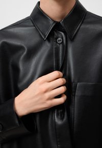 Schwarzes Lederhemd mit spitzem Kragen, vorderer Knopfverschluss und Brusttasche. Glatte Textur mit glänzendem Finish.