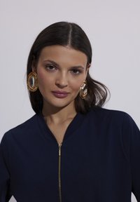 Granatowa kurtka zapinana na zamek z kołnierzykiem, z złotym zamkiem błyskawicznym. Duże złote okrągłe kolczyki z lustrującym środkiem. Gładka tekstura materiału.