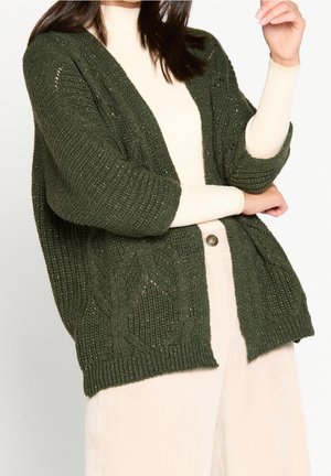 Femme portant un cardigan en tricot vert foncé sur un col roulé crème et un pantalon crème, bras croisés et main légèrement levée.