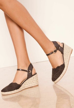 Beine in braunen Stoff-Wedge-Espadrilles mit Knöchelriemen und beigen geflochtenen Jutesohlen auf einem reflektierenden Boden.