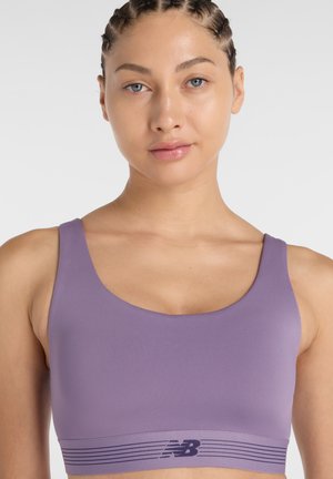 Jonge vrouw met gevlochten haar draagt een paarse sportbeha met het "NB"-logo, kijkt recht vooruit tegen een effen lichte achtergrond.