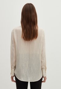 Camicia a maniche lunghe beige chiaro con un sottile motivo verticale e dettagli lucidi. Orlo arrotondato, polsini con bottoni e un tessuto morbido e testurizzato.