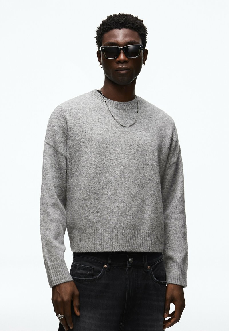 Homme portant des lunettes de soleil noires, un pull en maille gris, un jean noir, un collier chaîne en argent et des boucles d’oreilles créoles, debout sur un fond blanc.