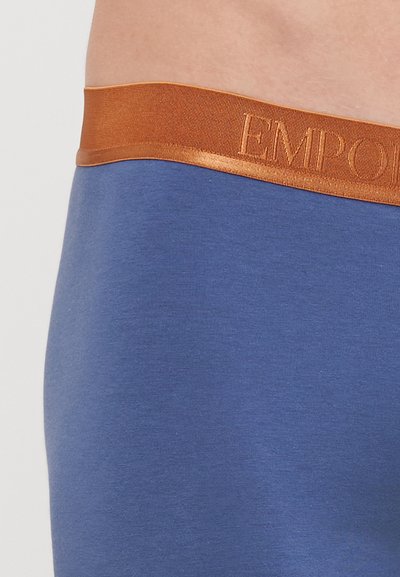 Modra tkaninska torba z gladko teksturo in širokim elastičnim pasom v zlato-oranžni barvi, na katerem je odtisnjen napis "EMPORIO."