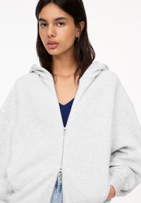 Hellegrauer Reißverschluss-Hoodie mit lockerer Passform und Kapuze. Verfügt über einen silbernen Reißverschluss, gerippte Bündchen und wird über einem dunkelblauen Oberteil getragen.
